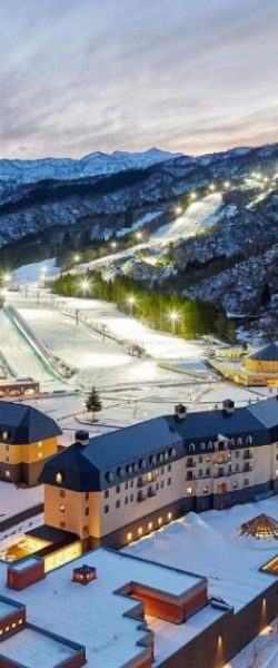 Snowy mountain ski resort at dusk with illuminated slopes and buildings in the foreground.